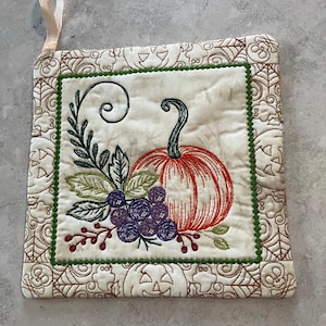 May include: A square quilted potholder featuring an embroidered orange pumpkin, purple berries, and green leaves. The border is decorated with brown stitched jack-o'-lanterns and spiderwebs. A light-colored fabric loop is attached to the top corner.