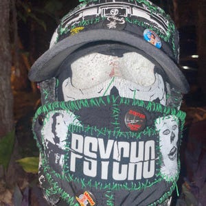 Könnte beinhalten: Eine schwarze und grüne Gesichtsmaske mit dem Wort "PSYCHO" darauf gestickt. Die Maske hat mehrere Patches und Pins, darunter einen Pin mit der Aufschrift "Exiter".