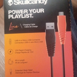 Może przedstawiać: Czarno-pomarańczowe opakowanie kabla ładującego Skullcandy. Opakowanie zawiera tekst &bdquo;POWER YOUR PLAYLIST&rdquo; oraz szczeg&oacute;ły dotyczące złącza USB-A do Micro-USB, szybkiego ładowania 12 W i długości 1,22 m.