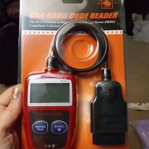 Może przedstawiać: Czerwono-czarny czytnik kodów OBDII z ekranem, przyciskami oznaczonymi jako "Enter/Exit" i "Scroll" oraz tekstem "OBDII/EOBD" z przodu. Czytnik kodów jest zapakowany w przezroczysty plastikowy blister z pomarańczowym tłem i tekstem "CAN OBDII CODE READER" i "Newer OBDII".