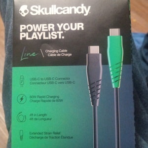 Może przedstawiać: Czarno-zielone pudełko kabla ładującego Skullcandy. Na pudełku widnieje napis "POWER YOUR PLAYLIST." oraz szczeg&oacute;ły dotyczące złącza USB-C do USB-C, szybkiego ładowania 60W i długości 1,2 m.