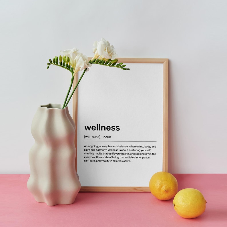 May include: A framed print with a white background and black text defining the word "wellness". The print is sitting on a pink surface with a vase of white flowers and lemons.