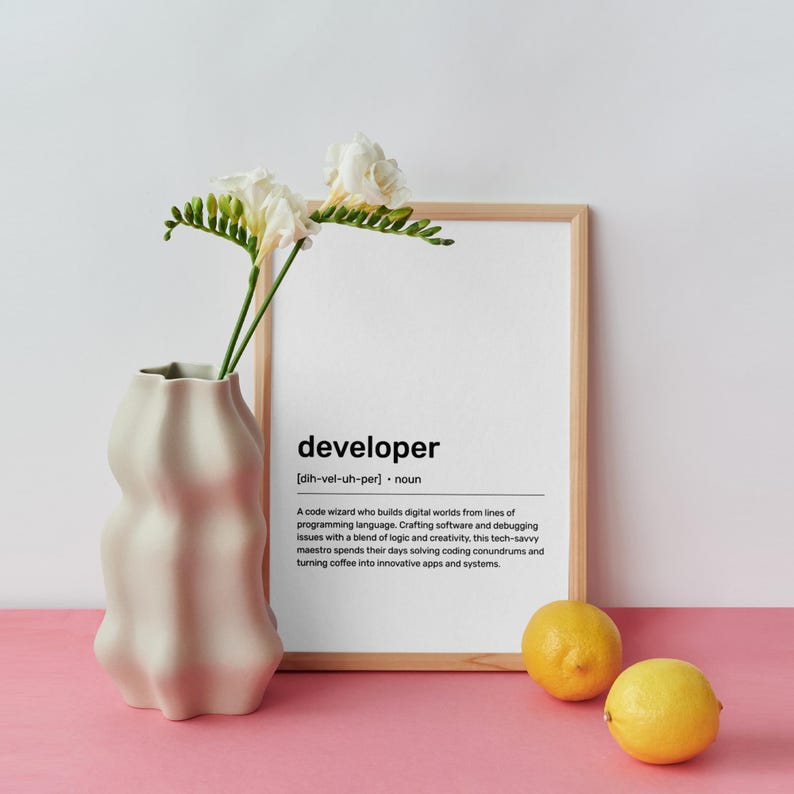 May include: A beige, wavy vase contains white flowers with green stems. A framed print with the definition of "developer" and two yellow lemons are on a pink surface.