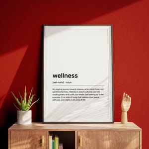 May include: A black and white printable poster with the word "wellness" defined as an ongoing journey towards balance where mind, body, and spirit find harmony. The poster is framed and hanging on a red wall.