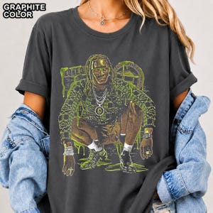 May include: A graphite-coloured t-shirt featuring a graphic print of a person with dreadlocks in a crouching pose. The print includes lime green accents, the text "ZONE 3", and a circular emblem. The shirt has short sleeves and a crew neckline.
