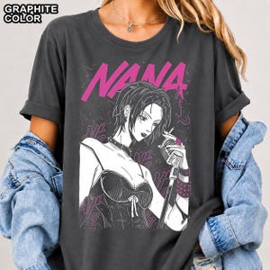 May include: A graphite-colored t-shirt featuring a graphic print of a character with short hair, a choker, and a corset top, holding a microphone. The text NANA is printed in pink above the character, and the words GRAPHITE COLOR appear in the top left.