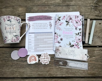 May include: A white ceramic mug, a Bible reading checklist, a floral notebook, a black pen, two markers, two circular discs, two stickers with religious text, a rectangular bookmark, and a clear plastic ruler with inch markings.
