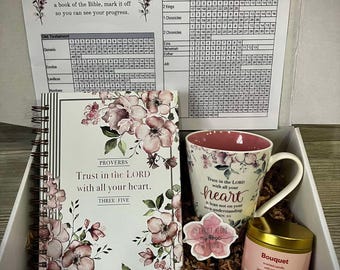 May include: A gift box filled with items for a Christian woman. The box contains a floral patterned journal, a mug with a floral design and the text "Trust in the Lord with all your heart", a floral patterned bookmark with the text "I am with you always", a pink heart-shaped object, a sticker with the text "In Christ Alone my hope is found", and a small jar of candle with the text "Bouquet".
