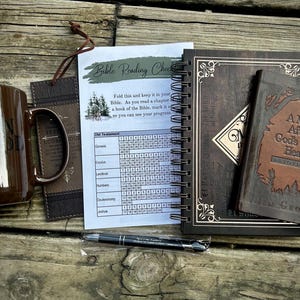 May include: A brown ceramic mug, a dark brown bookmark with a leather cord, a white Bible reading checklist paper, a black spiral-bound notebook with gold accents, a brown devotional book titled A Man After God's Own Heart, and a black ballpoint pen.