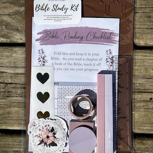 May include: A Bible study kit in a clear plastic sleeve containing a Bible reading checklist, a sheet of heart stickers, a floral sticker with text, two pink pens, a roll of washi tape, a round sticky note, and a 6-inch clear plastic ruler.