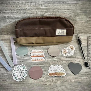 May include: A brown pencil case with the word "Silas" on it. The case is open and there are several stickers inside, including one that says "Jesus is worth everything you're afraid of losing."