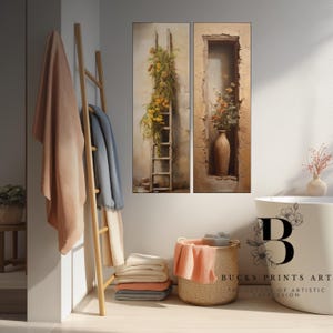 May include: Two framed wall art pieces. One depicts a wooden ladder adorned with yellow flowers. The other shows a vase with flowers in a stone alcove. A wooden towel rack holds blankets. A basket and a stack of folded blankets are in the foreground.