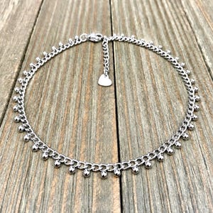 May include: A silver-toned chain necklace featuring a series of small, evenly spaced spherical beads dangling from the links. The necklace includes an adjustable chain extension finished with a small, flat heart-shaped charm at the end of the clasp.