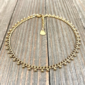 Puede incluir: Un collar de cadena en tono dorado que presenta una serie de pequeñas cuentas esféricas espaciadas uniformemente a lo largo del borde exterior de los eslabones. El collar incluye una cadena extensora ajustable con un pequeño dije de corazón.