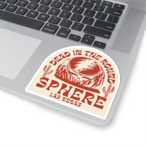 Pode incluir: Um adesivo vermelho e branco com o texto "Dead & Company" e "Sphere Las Vegas" em uma fonte retrô. O adesivo apresenta um gráfico de uma caveira com raios e um cacto.