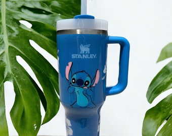 Könnte beinhalten: Blaue Reise-Thermoskanne aus Edelstahl mit blauem Griff und transparentem Deckel. Die Thermoskanne zeigt eine Zeichentrickfigur von Stitch aus Lilo & Stitch, weiße Hibiskusblüten und das Stanley-Logo.