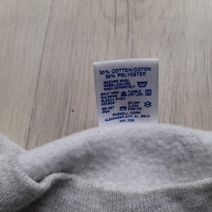 May include: A white tag with blue text and symbols detailing the care instructions for a garment. The tag reads "50% Cotton/Coton 50% Polyester", "Machine Wash Warm-Colors Wash Separately", "Only Non-Chlorine Bleach When Needed", "Tumble Dry Low", "Or Dry Clean Emblem", "Do Not Iron", "Russell Corp. Alexander City, AL 35010 WPL 7232".