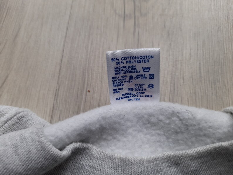 May include: A white tag with blue text and symbols detailing the care instructions for a garment. The tag reads "50% Cotton/Coton 50% Polyester", "Machine Wash Warm-Colors Wash Separately", "Only Non-Chlorine Bleach When Needed", "Tumble Dry Low", "Or Dry Clean Emblem", "Do Not Iron", "Russell Corp. Alexander City, AL 35010 WPL 7232".