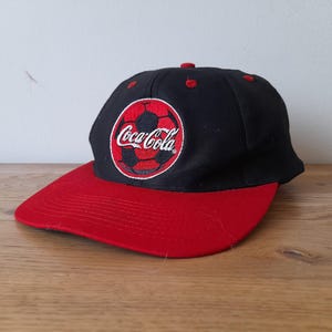 Könnte beinhalten: Eine schwarze und rote Baseballkappe mit einem Coca-Cola-Logo, das einen Fußball zeigt.