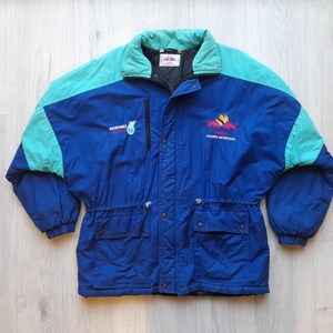 Puede incluir: Chaqueta azul y turquesa vintage con el logotipo y el texto de Petronas. La chaqueta tiene un cuello alto, cierre de cremallera y dos bolsillos delanteros. Las mangas y la parte superior de la espalda son turquesas, mientras que el cuerpo es azul.