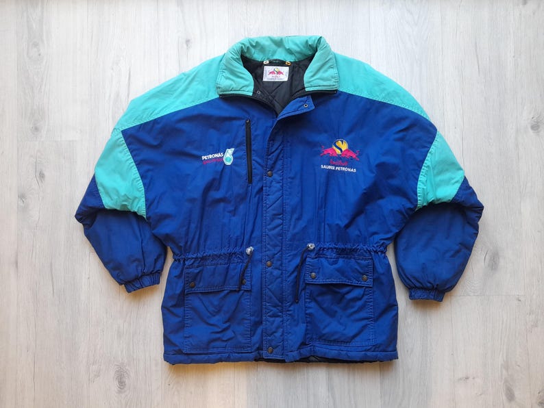 Puede incluir: Chaqueta azul y turquesa vintage con el logotipo y el texto de Petronas. La chaqueta tiene un cuello alto, cierre de cremallera y dos bolsillos delanteros. Las mangas y la parte superior de la espalda son turquesas, mientras que el cuerpo es azul.