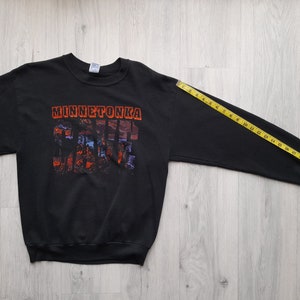 May include: Black long-sleeve sweatshirt with a graphic print of "Minnetonka" and a scenic image. The sweatshirt is laid flat on a wooden surface with a measuring tape.