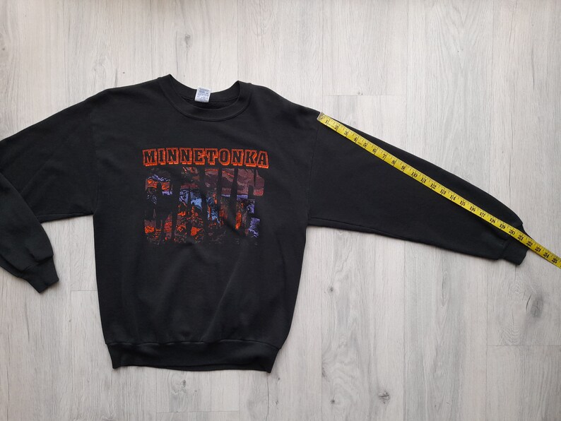 May include: Black long-sleeve sweatshirt with a graphic print of "Minnetonka" and a scenic image. The sweatshirt is laid flat on a wooden surface with a measuring tape.