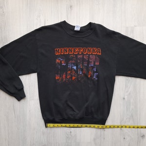 May include: Black long-sleeve sweatshirt with a graphic print of the text "Minnetonka" and a scenic landscape. The sweatshirt has a crew neck and ribbed cuffs.