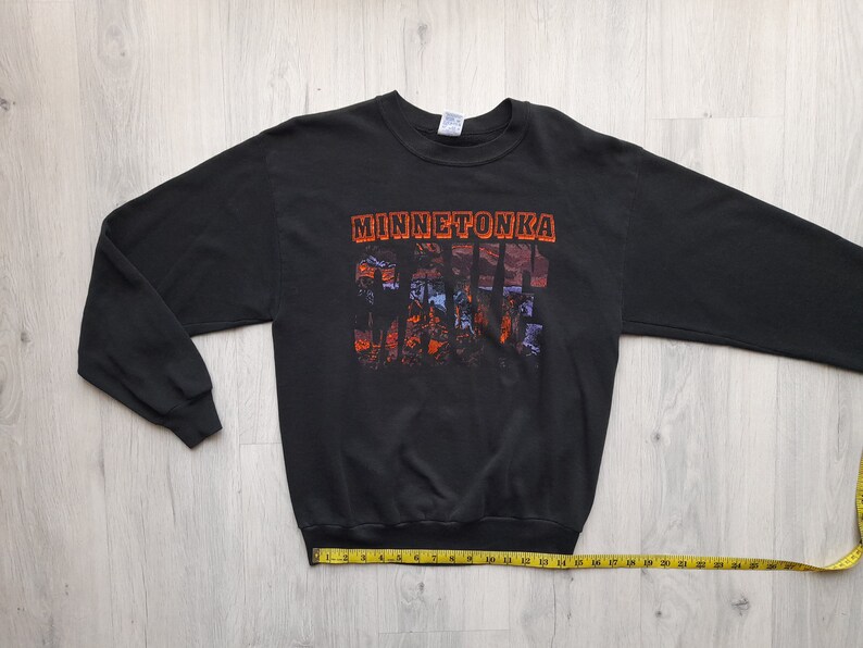 May include: Black long-sleeve sweatshirt with a graphic print of the text "Minnetonka" and a scenic landscape. The sweatshirt has a crew neck and ribbed cuffs.