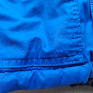 Puede incluir: Primer plano de una secci&oacute;n de una chaqueta azul brillante. La imagen destaca la textura de la tela y los detalles de las costuras. La chaqueta parece estar hecha de un material duradero, posiblemente resistente al agua, con costuras visibles y un dise&ntilde;o acolchado.