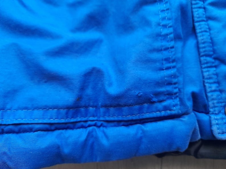 Puede incluir: Primer plano de una secci&oacute;n de una chaqueta azul brillante. La imagen destaca la textura de la tela y los detalles de las costuras. La chaqueta parece estar hecha de un material duradero, posiblemente resistente al agua, con costuras visibles y un dise&ntilde;o acolchado.