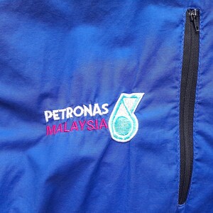 Puede incluir: Primer plano de una chaqueta azul con una cremallera negra y el logotipo de Petronas Malaysia bordado en la parte delantera. El logotipo es un emblema blanco, turquesa y rosa.