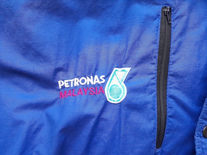 Puede incluir: Primer plano de una chaqueta azul con una cremallera negra y el logotipo de Petronas Malaysia bordado en la parte delantera. El logotipo es un emblema blanco, turquesa y rosa.