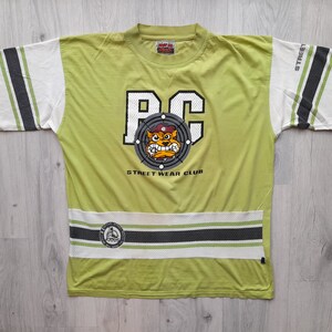 May include: A lime green t-shirt with a white and black striped design on the sleeves and bottom hem. The shirt features a large graphic of a cartoon dog wearing a red hat and a target with the letters "PC" in the center. The text "Street Wear Club" is printed below the graphic.