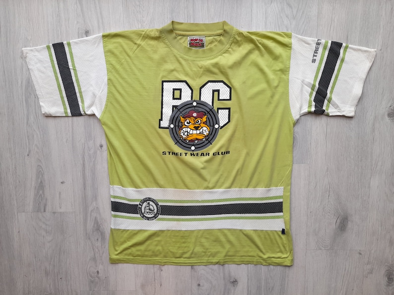 May include: A lime green t-shirt with a white and black striped design on the sleeves and bottom hem. The shirt features a large graphic of a cartoon dog wearing a red hat and a target with the letters "PC" in the center. The text "Street Wear Club" is printed below the graphic.