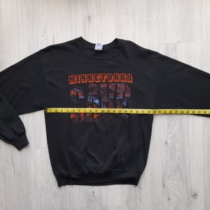 May include: Black long-sleeve sweatshirt with a graphic print of "Minnetonka" in orange and red letters. The graphic is made up of small images of buildings and trees.