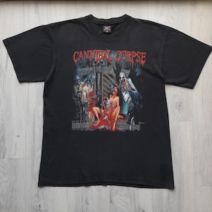 Puede incluir: Camiseta negra con el nombre de la banda "CANNIBAL CORPSE" en letras rojas que gotean sobre una ilustración gráfica. La ilustración representa figuras en una escena oscura de terror. Una advertencia "ADVISORY" está impresa en la parte inferior.