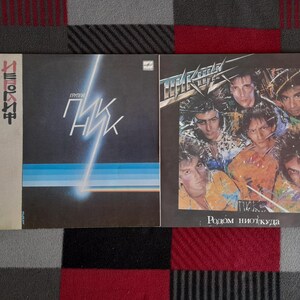 Peut inclure: Deux pochettes d'album vinyle. La pochette de gauche est sombre avec un design bleu et blanc et du texte cyrillique. La pochette de droite présente un groupe d'hommes aux cheveux longs et le texte cyrillique "Родом ніоткуда".