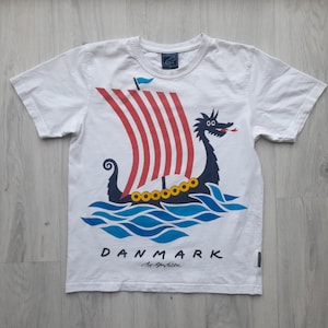 Könnte beinhalten: Weißes T-Shirt mit einem Wikingerschiff-Design. Das Schiff hat rot-weiß gestreifte Segel, eine blaue Flagge und eine Drachenfigur. Das Wort "DANMARK" ist darunter gedruckt. Das T-Shirt hat kurze Ärmel.
