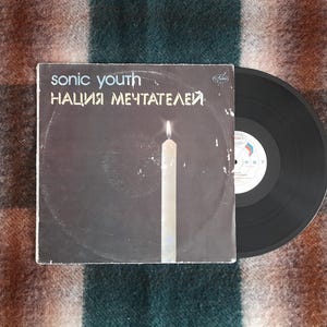 Może przedstawiać: Czarna płyta winylowa z białą etykietą. Okładka płyty jest czarna z białym tekstem, który brzmi "sonic youth" i "НАЛИЯ МЕЧТАТЕЛЕЙ". Biała świeca znajduje się w środku okładki.