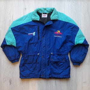 Puede incluir: Chaqueta vintage azul y turquesa con cuello alto y cintura ajustable. La chaqueta presenta el logotipo de Petronas y un emblema colorido. Tiene dos bolsillos delanteros y una cremallera negra.