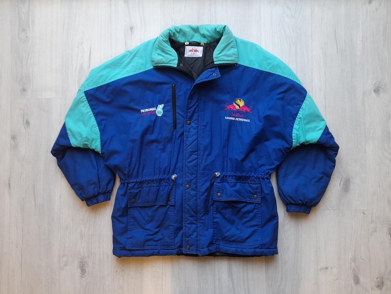 Puede incluir: Chaqueta vintage azul y turquesa con cuello alto y cintura ajustable. La chaqueta presenta el logotipo de Petronas y un emblema colorido. Tiene dos bolsillos delanteros y una cremallera negra.