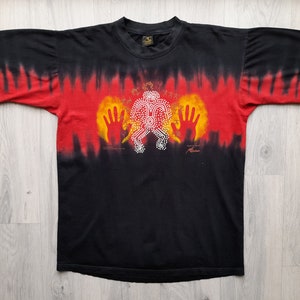 May include: Black tie-dye t-shirt with a red, yellow, and white Aboriginal art design. The design features a figure with outstretched hands and a pattern of dots.