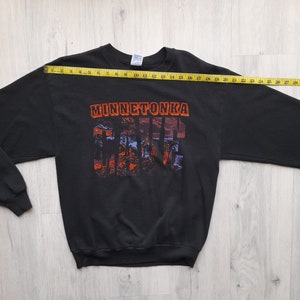 May include: Black long-sleeve sweatshirt with a graphic print of the text "Minnetonka" and a colorful landscape scene. The sweatshirt is laid flat on a wooden surface with a measuring tape.