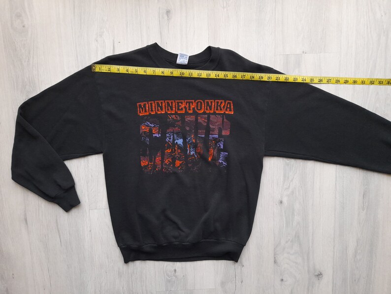 May include: Black long-sleeve sweatshirt with a graphic print of the text "Minnetonka" and a colorful landscape scene. The sweatshirt is laid flat on a wooden surface with a measuring tape.