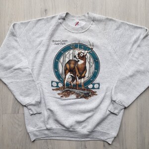 May include: Gray sweatshirt with a graphic of a white-tailed deer in a forest setting. The graphic includes the text "Brown County State Park".