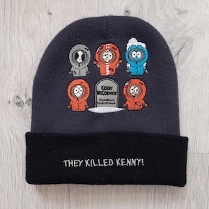 May include: A dark grey beanie hat with a black brim, embroidered with cartoon characters in various outfits. The text "THEY KILLED KENNY!" is displayed on the brim. The hat has a playful, cartoon-inspired design.