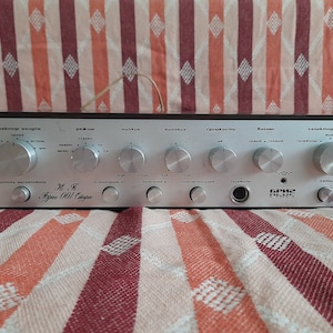 Op de afbeelding: Een zilveren en zwarte stereo versterker met meerdere knoppen en draaiknoppen. De versterker heeft de tekst "Hi-Fi "Brigadir 001" Stereo" aan de voorkant.