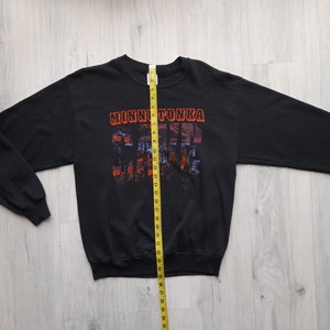 May include: Black long-sleeve sweatshirt with a graphic print of a sunset scene and the text "MINNETONKA" in orange and red. The sweatshirt is laid flat on a wooden surface with a measuring tape beside it.