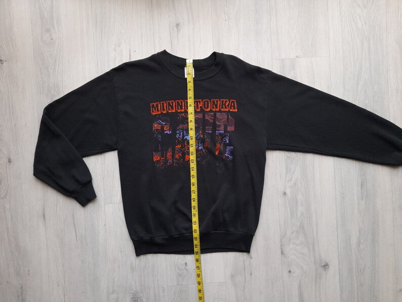 May include: Black long-sleeve sweatshirt with a graphic print of a sunset scene and the text "MINNETONKA" in orange and red. The sweatshirt is laid flat on a wooden surface with a measuring tape beside it.